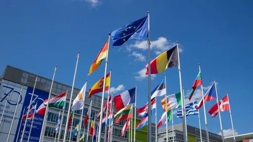 Flags of various countries flying in front of a modern building under a blue sky.